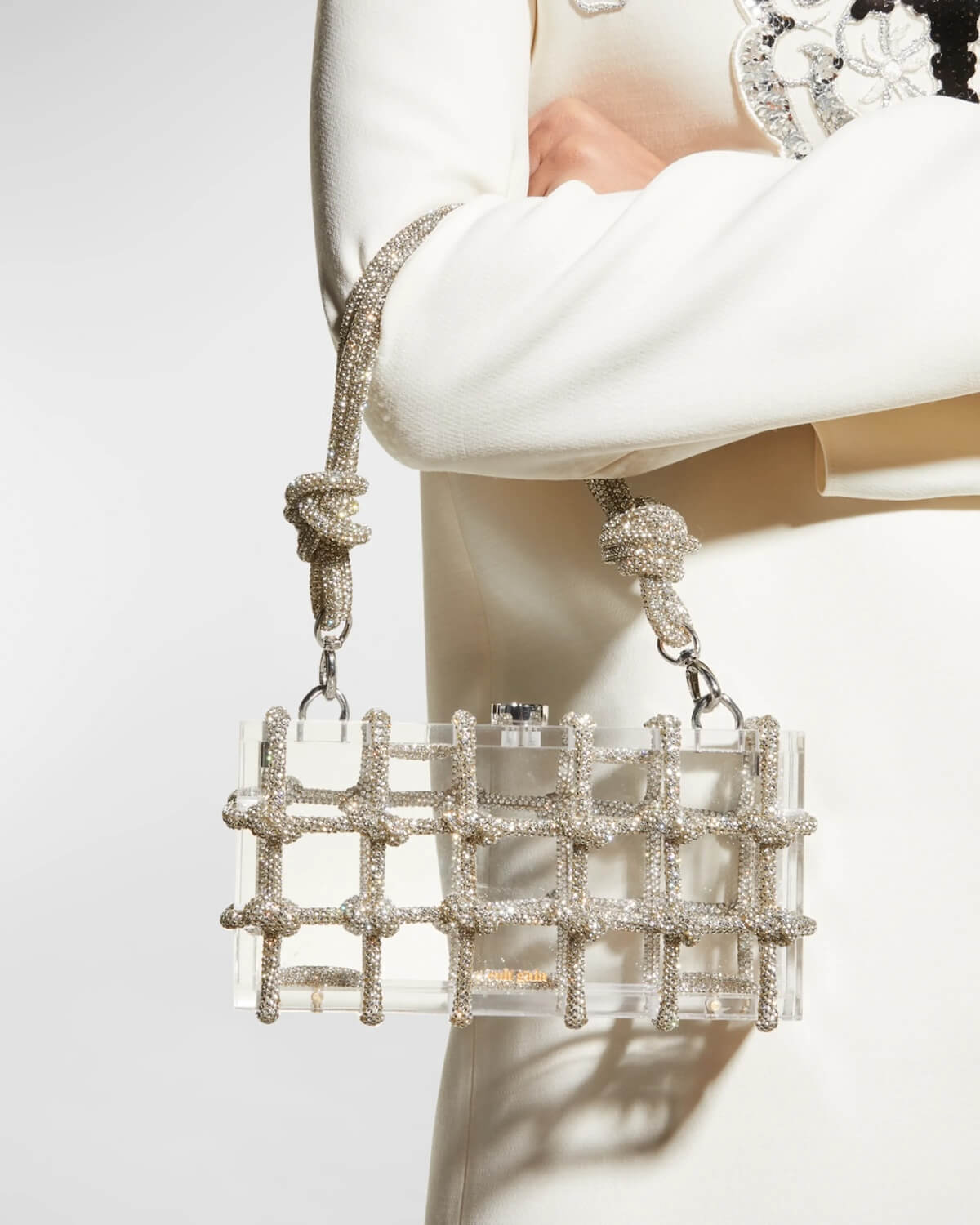 A person in white holds a clear rectangular handbag with diamond-encrusted rope details and two knotted rope handles.
