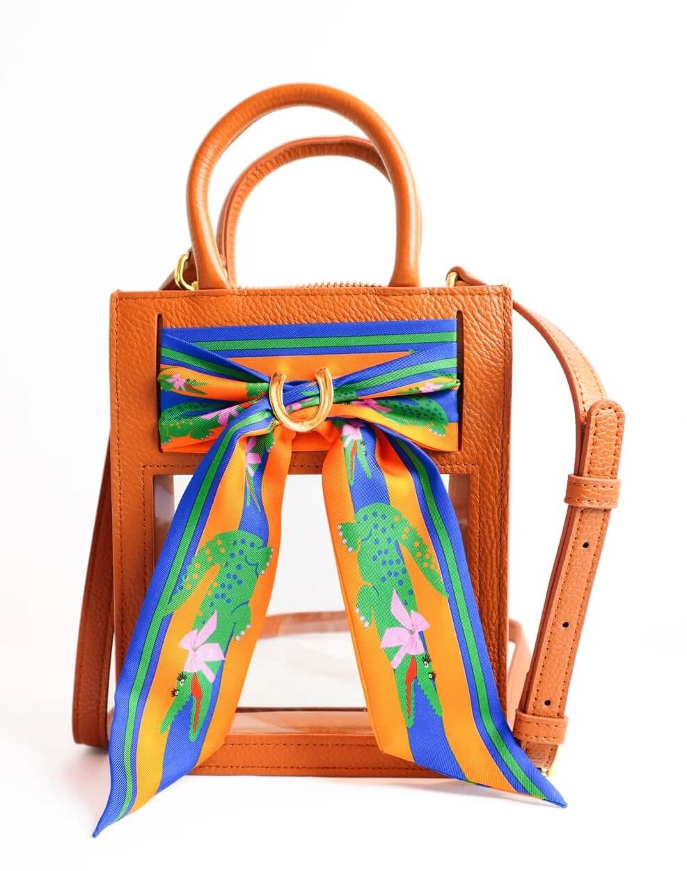 A small, rectangular, brown leather handbag with a detachable shoulder strap and double handles. The front is accented with an orange, blue, and green patterned scarf tied in a bow. This stylish piece combines the charm of vintage elegance while offering the modern versatility of a clear bag.