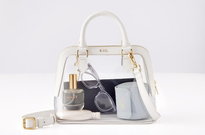 Clear bag with gold details containing a perfume bottle, sunglasses case, small blue bag, and white accessory on a white background. This chic paste-over look effortlessly combines sophistication and functionality.