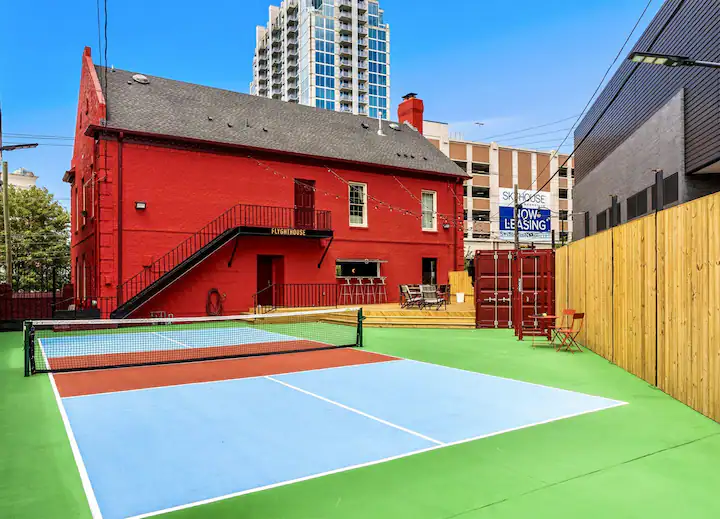 An outdoor area features a blue and green pickleball court near a bright red building with "Putnam House" painted on it, surrounded by wooden and metal fences, with city buildings in the background. This setup is truly one of the craziest Airbnbs you could stumble upon!