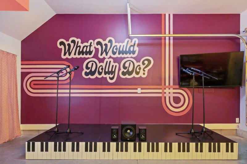 A small stage with a piano keyboard design on the floor. Two microphones on stands and a speaker are on the stage. The back wall features a sign that says "What Would Dolly Do?" and a mounted TV.