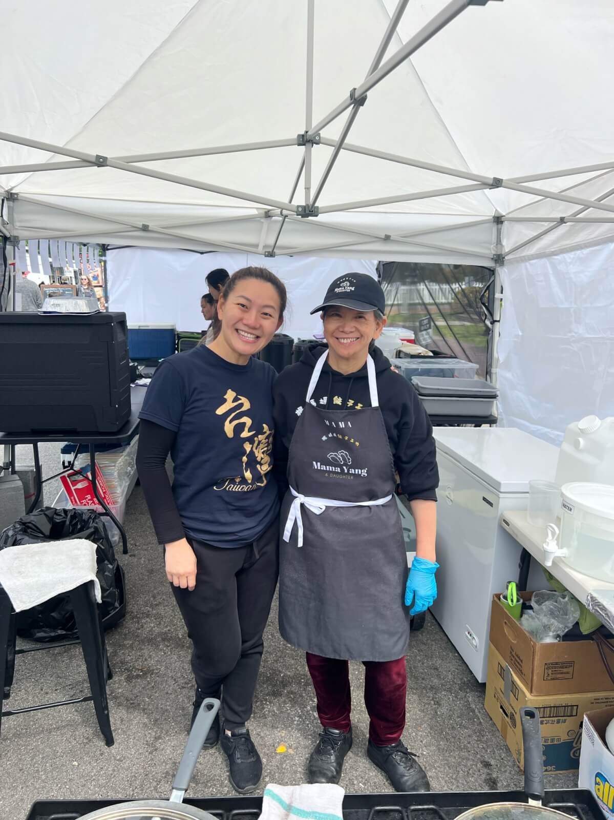 Two women, their FACES beaming with joy, stand under a white tent at Mama Yang's food stall. One wears a black t-shirt, while the other dons a black apron and cap, with disposable gloves. Cooking equipment and supplies surround them.