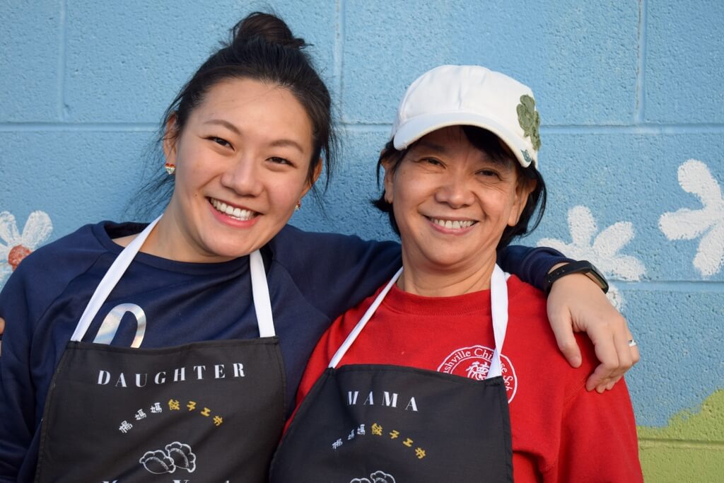 Two women stand side by side in front of a painted wall, both smiling and wearing aprons labeled "Daughter" and "Mama Yang." One woman is wearing a white cap, their happy faces reflecting their close bond.