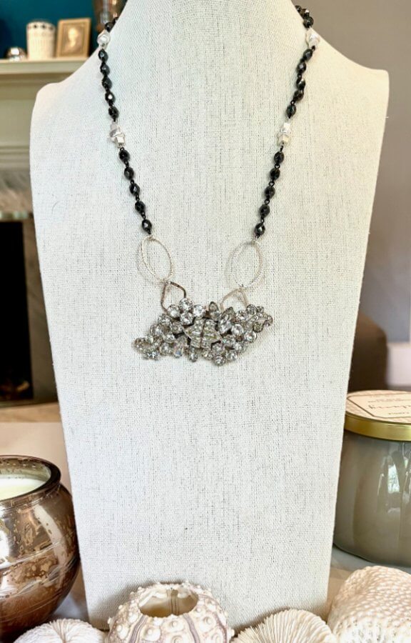 A beaded necklace with a large, intricate central pendant is displayed on a white bust, surrounded by decorative items on a table, exuding the unmistakable elegance of a Stacey Pierce-Nickle creation.