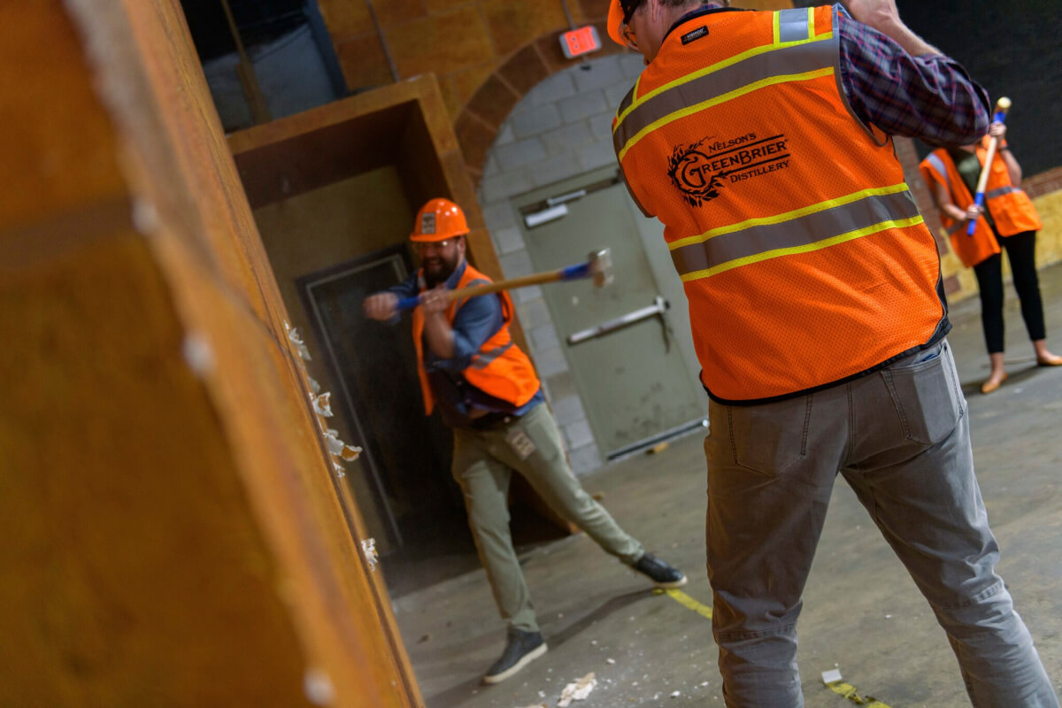 People in orange safety vests and hard hats use sledgehammers to demolish a wall in an indoor setting at Nelson's Greenbrier Distillery.