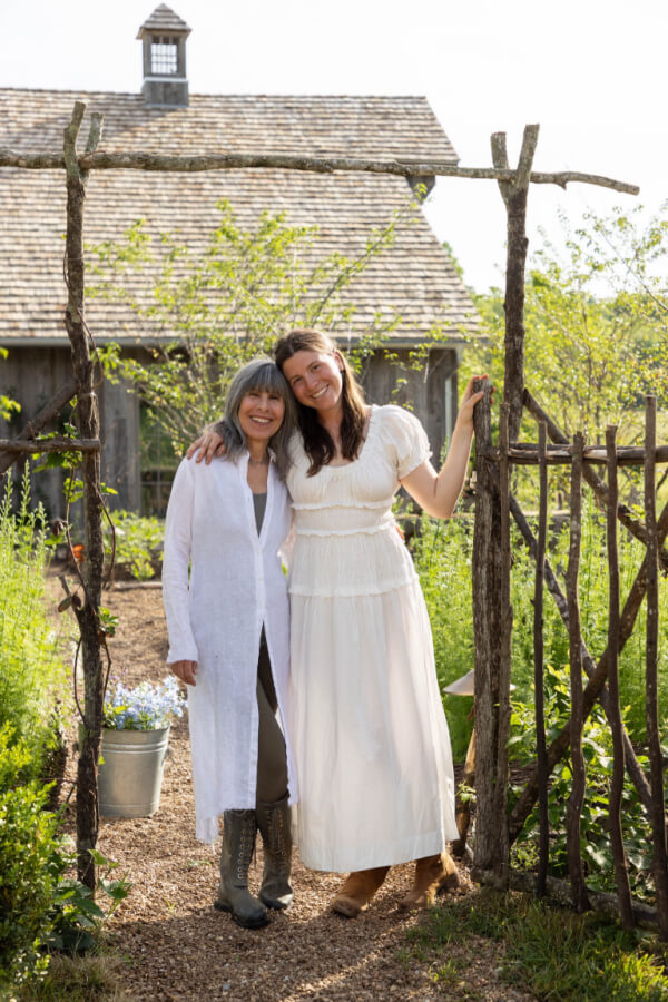 Two women stand together, smiling and posing at an open garden gate. An old wooden building and lush greenery serve as the backdrop for this charming gathering space. One woman wears a long white cardigan, while the other dazzles in a white dress.