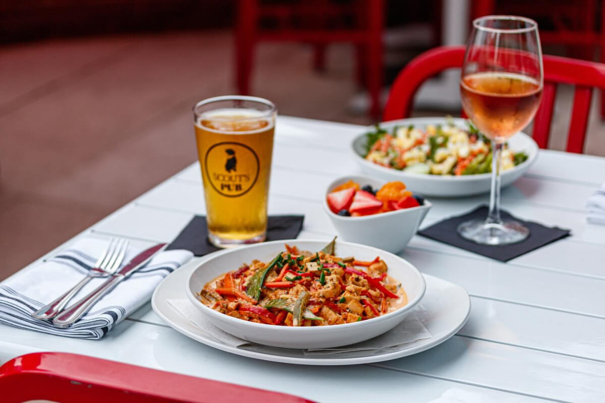 A white table with a plate of pasta, a glass of beer, a bowl of fruit, and a glass of wine. Another plate of salad is partially visible in the background. Red chairs surround the table.