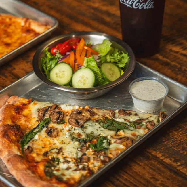 A metal tray holds a slice of vegetable pizza, a fresh salad with cucumbers, tomatoes, and carrots, a small cup of dressing, and a drink in a Coca-Cola cup, all on a wooden table.