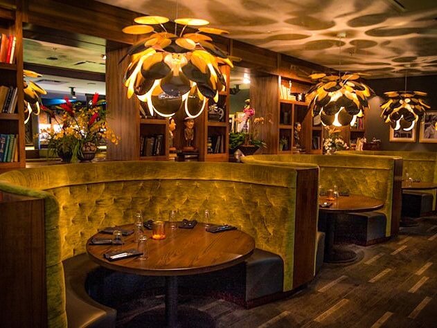 A cozy restaurant interior with round booths, wooden tables, and unique pendant lights creating a warm ambiance. Shelves with books and decorative items line the walls.