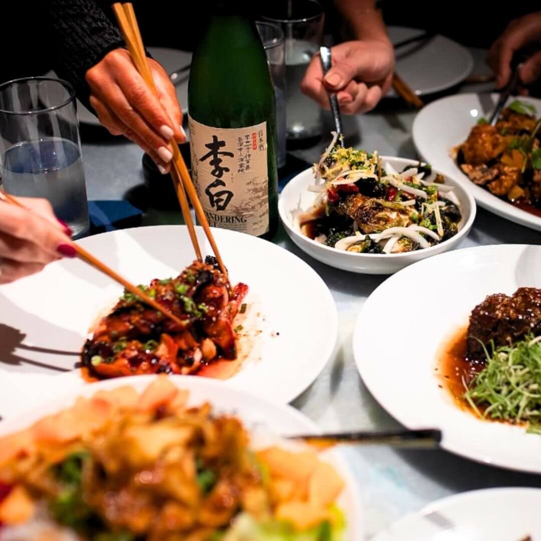 People using chopsticks to share various dishes at a restaurant, with a bottle of sake at the center.