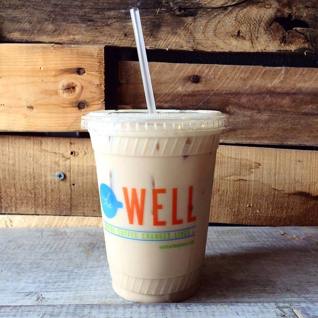 A plastic cup with a straw, filled with iced coffee, sits on a wooden surface against a rustic wood backdrop. The cup has a label that reads "The Well."
