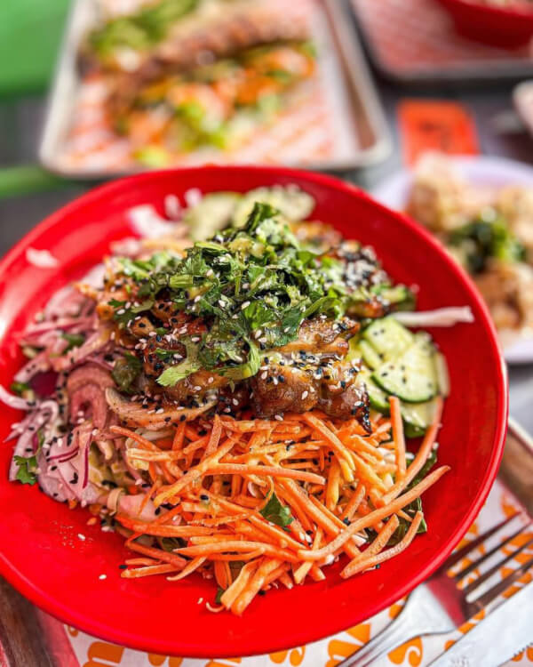 A close-up of a red bowl containing a colorful salad with shredded carrots, sliced cucumbers, pickled onions, greens, and sesame seeds. 