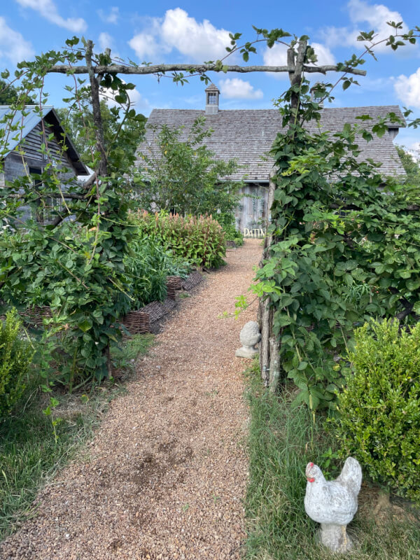 A gravel path leads through a trellis covered in greenery towards a rustic house, with various plants, garden decorations, and a white chicken statue on the right. Nearby sits The Shed, adding an extra touch of charm to the picturesque scene.