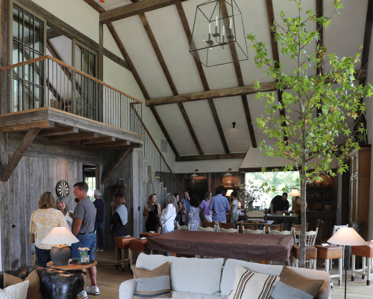 A group of people gather in The Shed's rustic living room, featuring high ceilings, wooden beams, and various seating arrangements. The cozy space includes a dartboard, a tree, and offers a charming view into the kitchen area.
