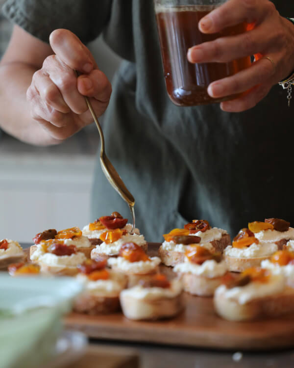 A person from The Shed drizzles honey onto crostini topped with various ingredients, using a spoon, while holding a jar of honey.