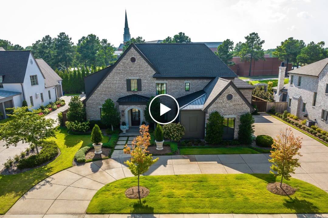 A brick house with a well-maintained lawn and driveway is nestled in the picturesque neighborhood of Chapel Woods, Germantown TN. Surrounded by green trees and other homes, it offers a charming view with a church steeple visible in the background.