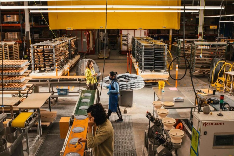 Workers in a ceramics factory operate machinery and handle products, surrounded by rows of stacked ceramic items and East Fork Pottery pieces alongside industrial equipment.