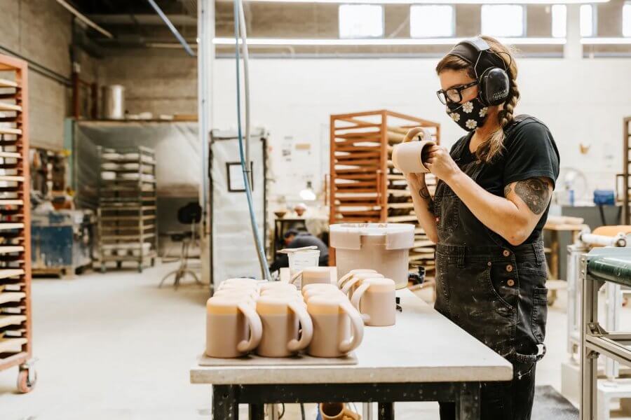 A person wearing a mask and ear protection inspects a ceramic mug in an East Fork Pottery workshop. A table with multiple mugs is in front of them, and shelves and equipment are visible in the background.