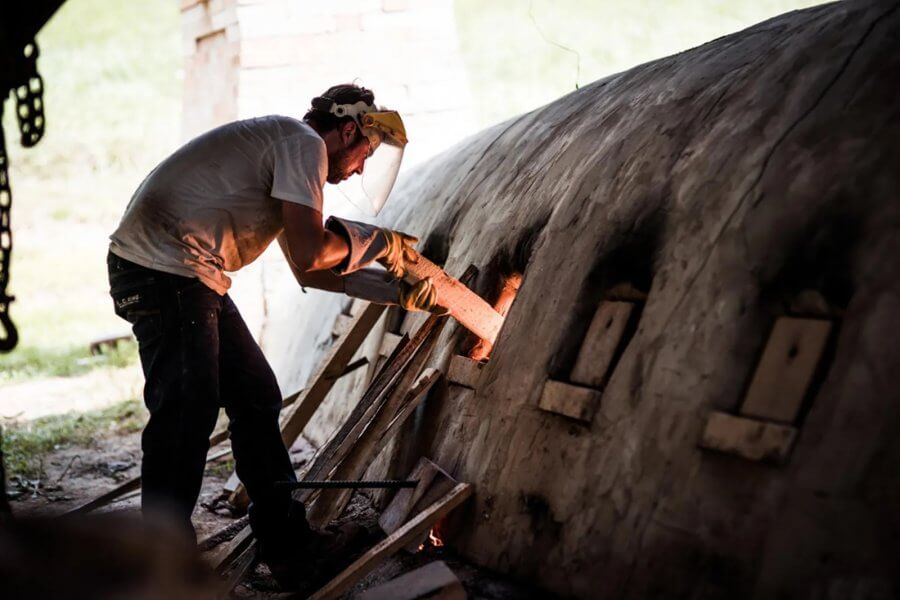A person in protective gear inserts wood into the openings of a large East Fork Pottery kiln.