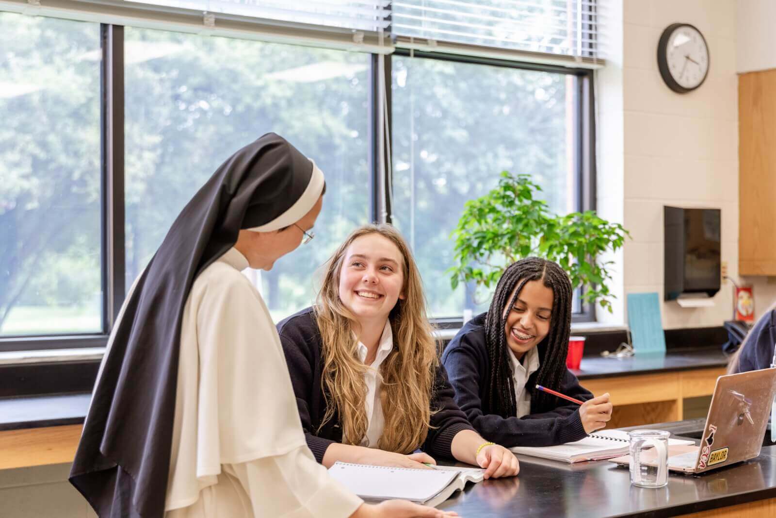 A smiling nun speaks with two students in a Nashville private school classroom. The students are seated at a table, working in notebooks. A clock and a potted plant are in the background.