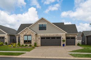 A single-story house with a stone facade and gray siding, located on Dressage Circle in Louisville, KY. Featuring three large garage doors, a well-maintained lawn, and a clear blue sky with some clouds in the background, this charming home has been standing proudly since 2007.
