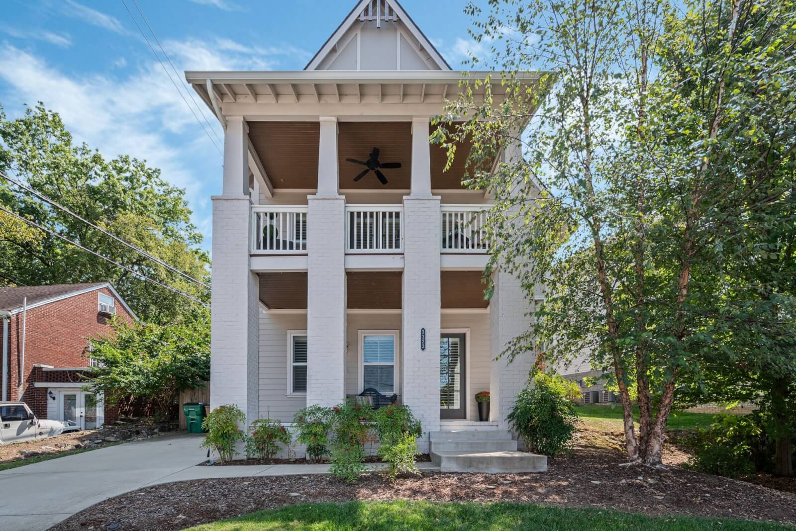 A front view of a two-story house on 3323 Orleans Dr, showcasing white brick columns and an upper-level porch. The yard features a tree and a small landscaped garden, with a glimpse of the neighboring house to the left.