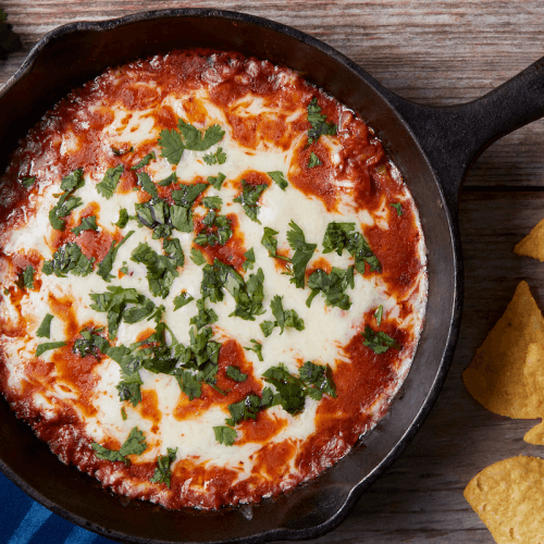 A cast iron skillet filled with baked melted cheese topped with diced cilantro, accompanied by tortilla chips on a wooden surface.