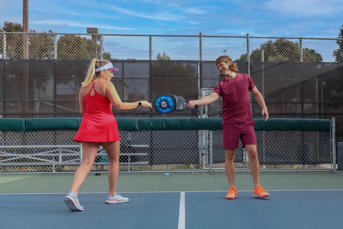 Two people on an outdoor Birmingham pickleball court tap paddles together, both wearing athletic attire and standing near the net.