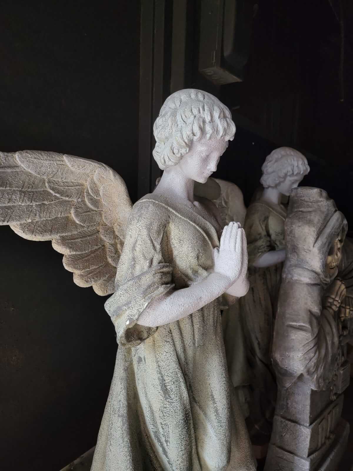 A stone angel statue with detailed wings and folded hands in a praying pose, set against a dark background at the Conjure Haunted Attraction.