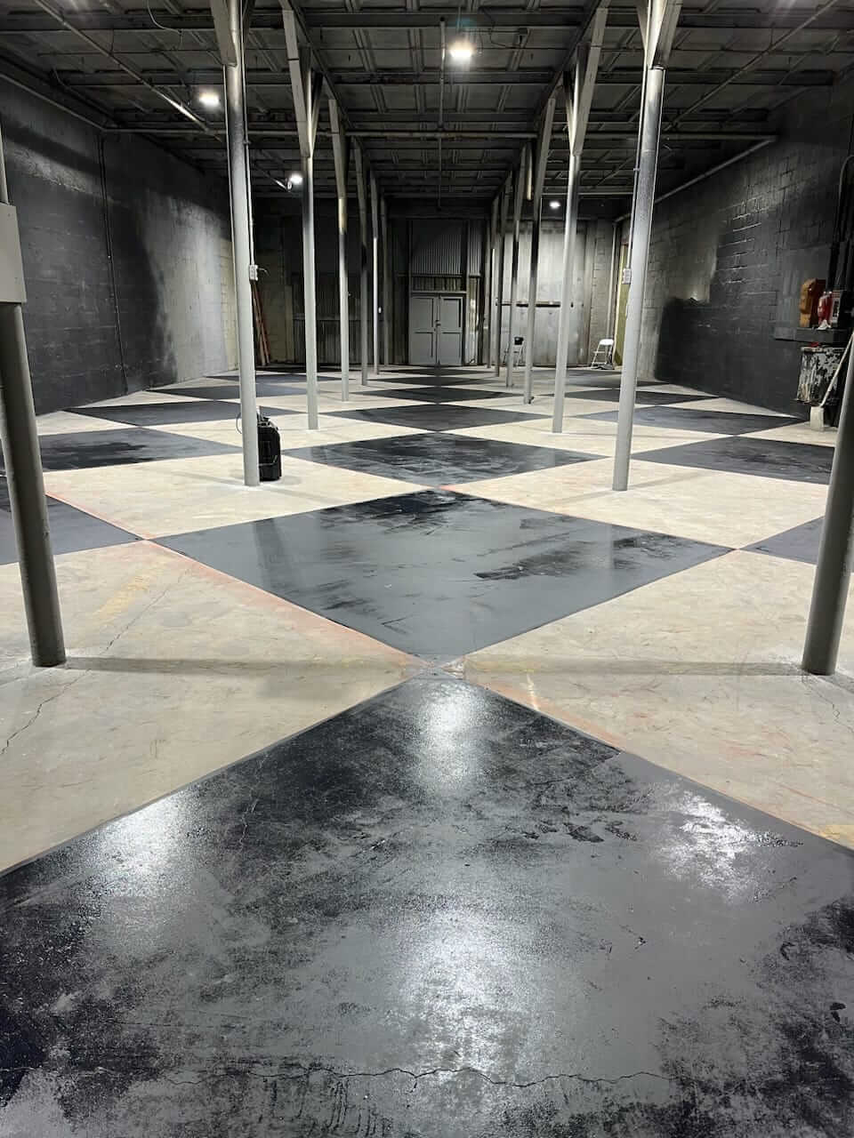 A warehouse interior with concrete floors painted in a black and white diamond pattern. Multiple columns support the ceiling, and various items are scattered in the background at the Conjure Haunted Attraction.