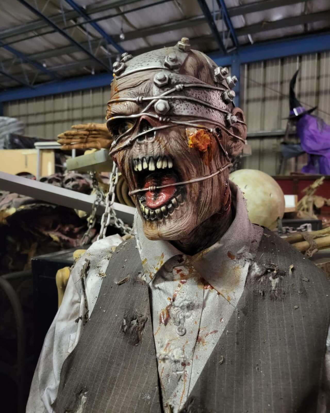 Close-up of a menacing Halloween prop with exposed teeth, an open mouth, and metal wires wrapping its head, dressed in a tattered vest and shirt at the Conjure Haunted Attraction