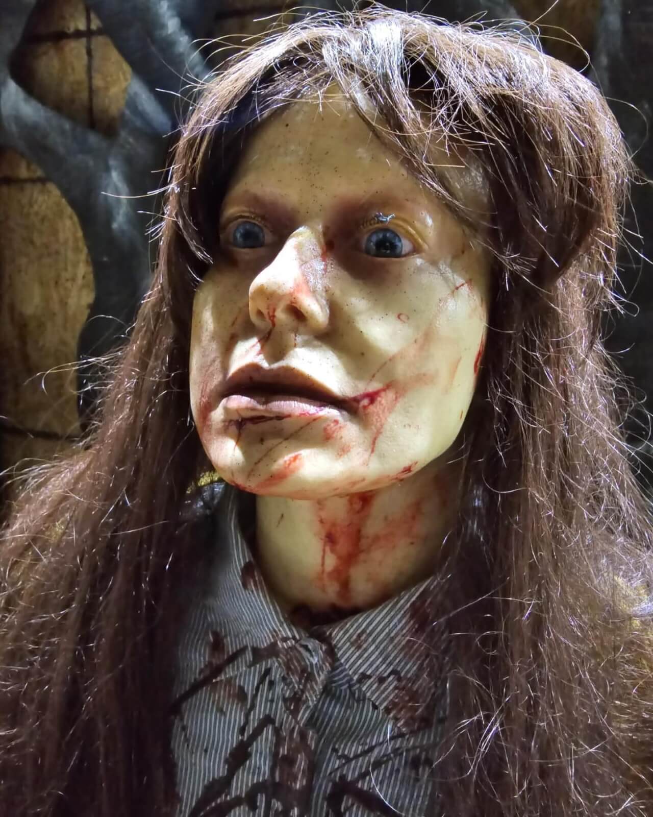 A close-up of a mannequin with a pale, blood-smeared face, long dark hair, and blue eyes, designed to look frightening at the Conjure Haunted Attraction.