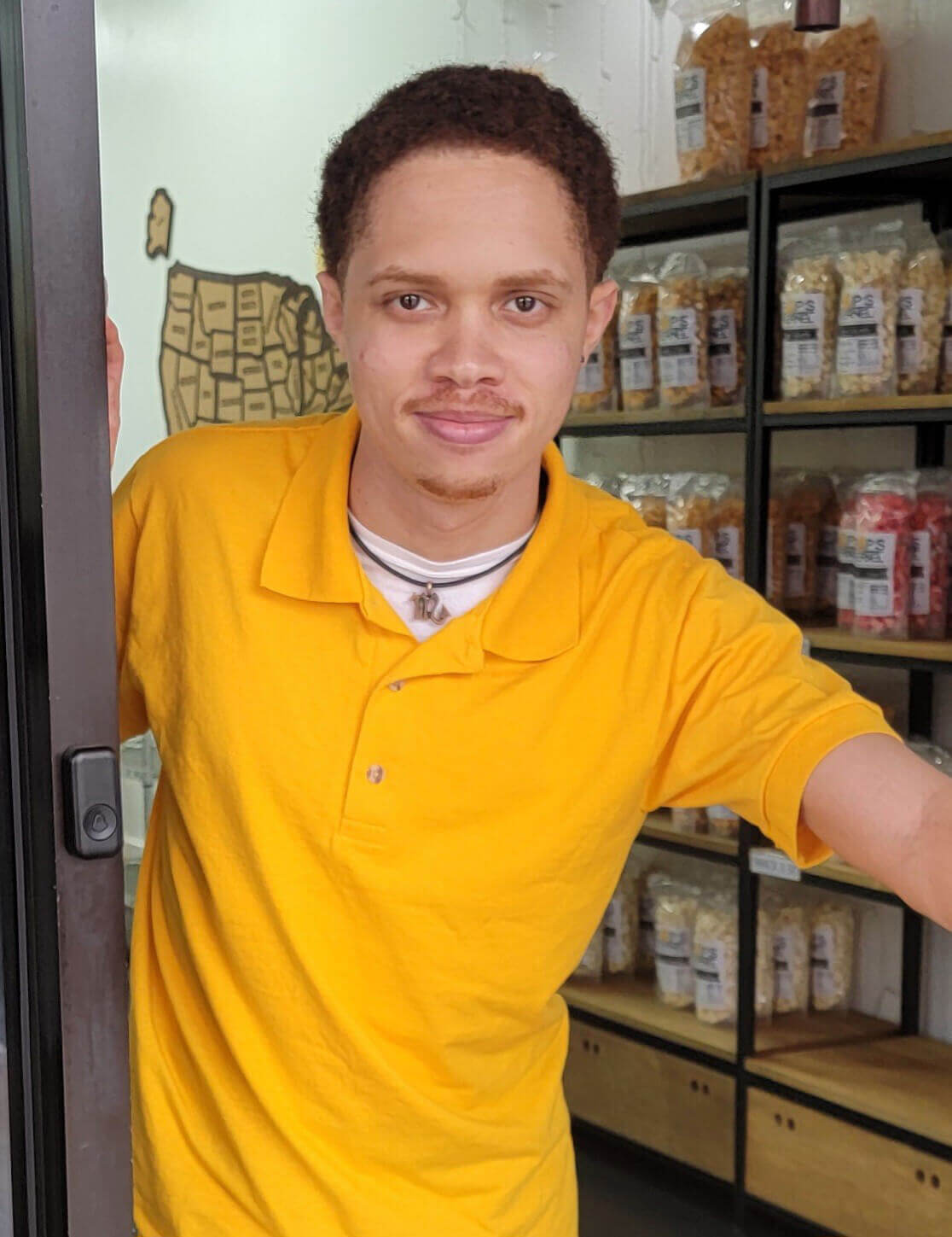 A man in a yellow shirt stands in a doorway, smiling. Behind him are shelves filled with various packaged goods, including Pop's Kernel popcorn.