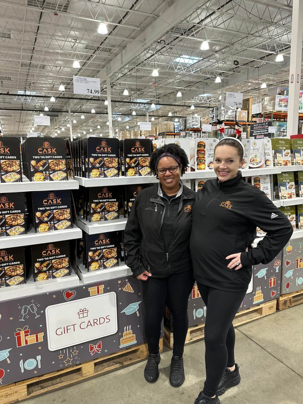 Two women dressed in black stand in front of a display of boxed food products at a retail store. A sign for gift cards is visible on the display.