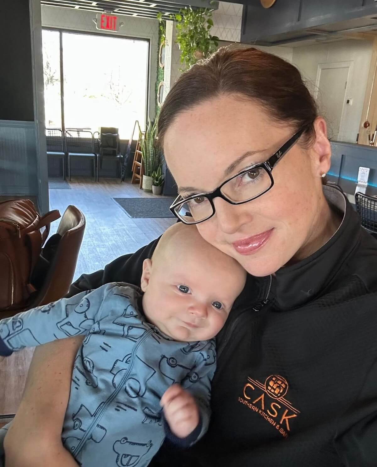 A woman with glasses, wearing a dark sweater, holds a baby dressed in blue with animal prints in a restaurant or cafe setting.