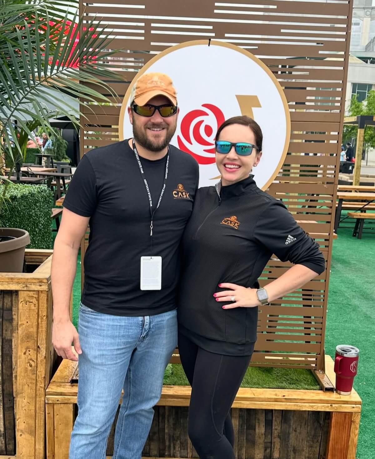 Two people in casual attire, both wearing sunglasses, stand together and smile in front of a wooden cask backdrop featuring a red rose logo.