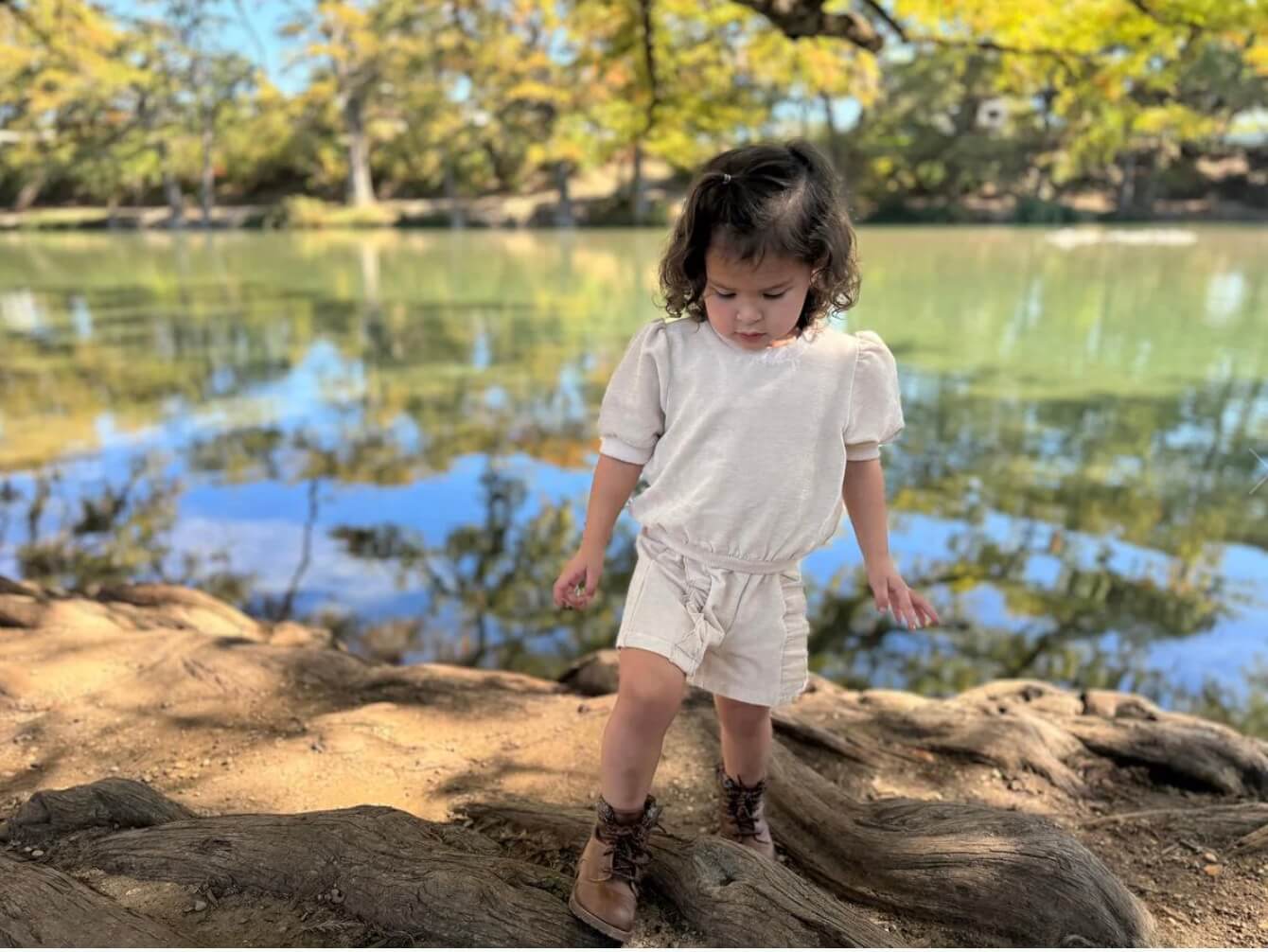 A young child wearing a white outfit and brown boots walks on a tree root by a calm lake with trees in the background.