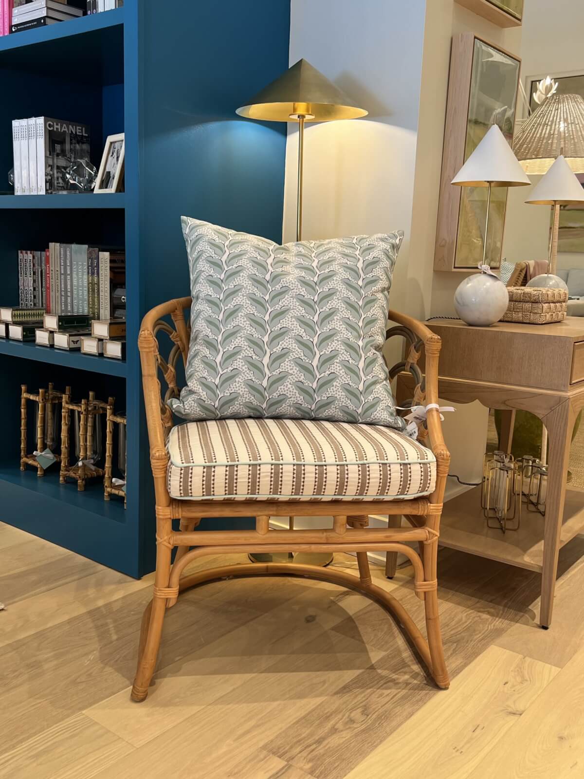 A rattan chair with a striped cushion and a patterned pillow is placed in front of a bookshelf and beside a wooden desk with decorative lamps. A brass floor lamp stands behind the chair.