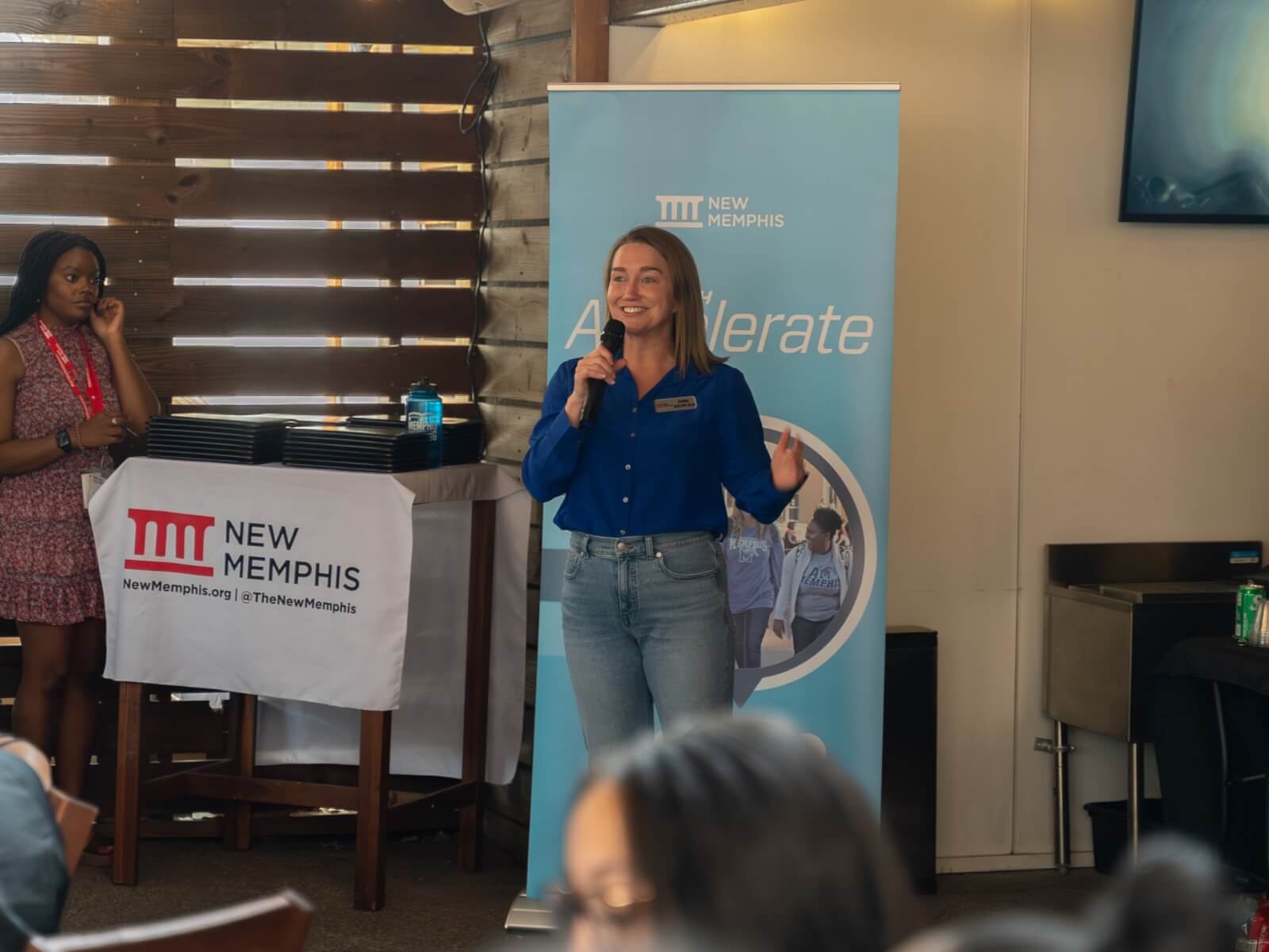 A woman in a blue shirt speaks into a microphone in front of a banner and a table spotlighting the New Memphis logo at an indoor event.
