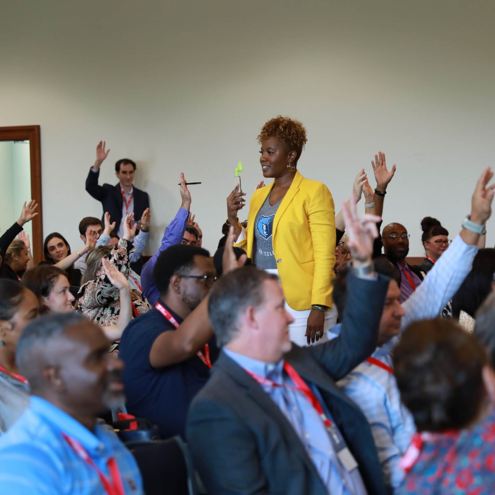 A woman in a yellow jacket stands holding a microphone in a classroom filled with seated individuals, many raising their hands.