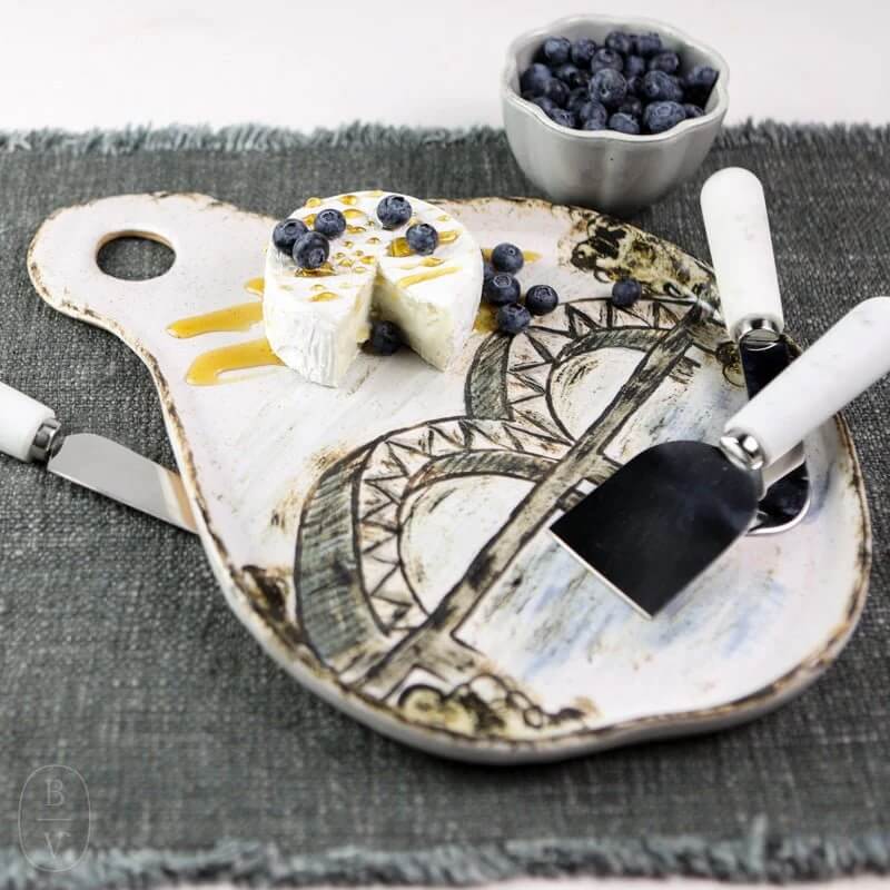 A ceramic cheese board with an image of a bridge holds brie cheese topped with blueberries and honey. A small bowl of blueberries and cheese utensils rest on a gray cloth