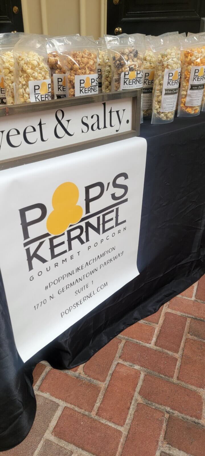 Display table with assorted popcorn bags from Pop's Kernel Gourmet Popcorn. A banner reads "Pop's Kernel Gourmet Popcorn" with a contact address and social media hashtag.
