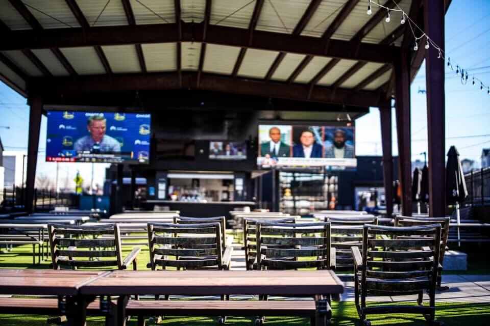 An outdoor seating area with multiple tables and chairs under a roof, featuring two large TV screens showing sports-related programming, is one of the great spots in Nashville to watch football.