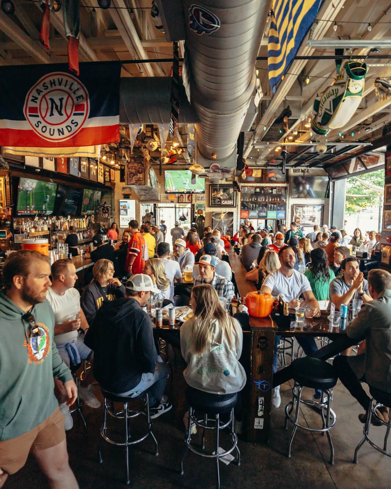 A bustling bar in Nashville filled with patrons watching football on multiple screens, decorated with sports memorabilia and flags hanging from the ceiling.