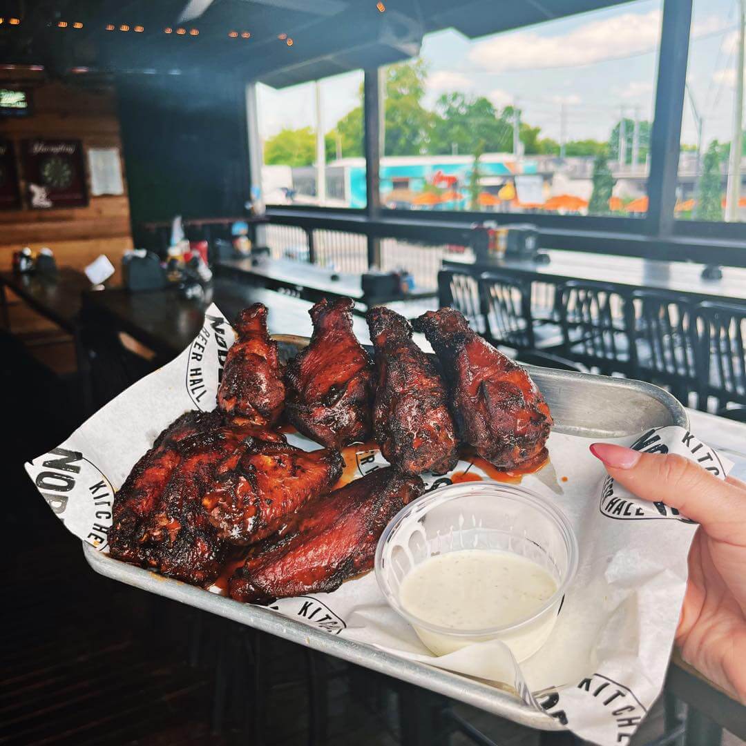 A tray of grilled chicken pieces beside a dipping sauce container held by a person in a restaurant with outdoor views—one of the great spots to watch football in Nashville.