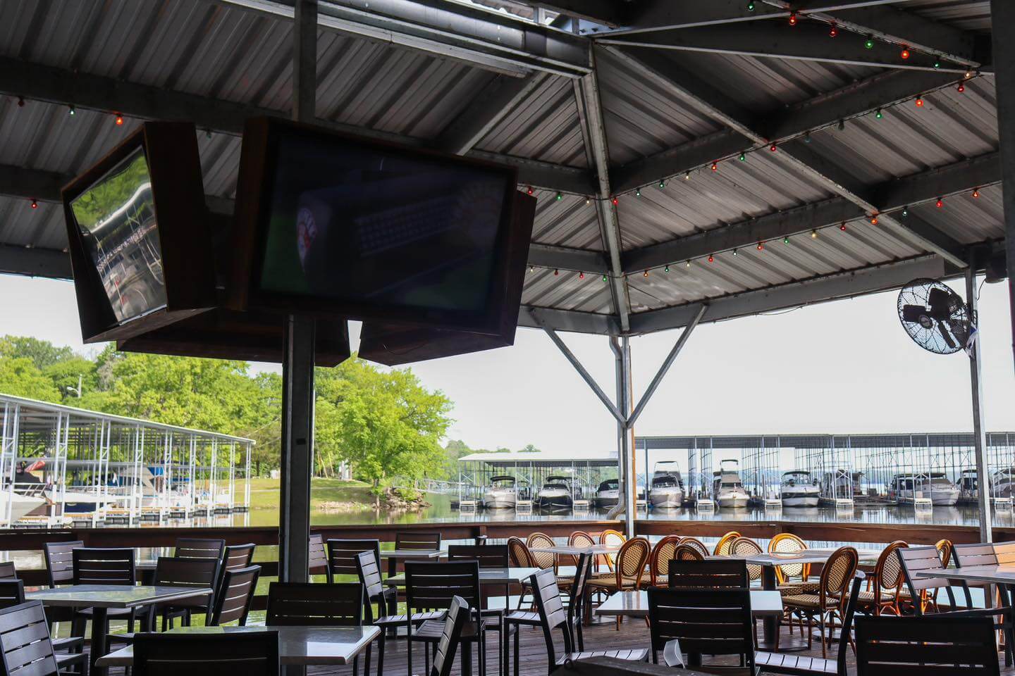 An outdoor, covered seating area with tables and chairs near a marina docked with boats. Spots to watch football on ceiling-mounted TVs and enjoy the lights hanging under the metal roof.