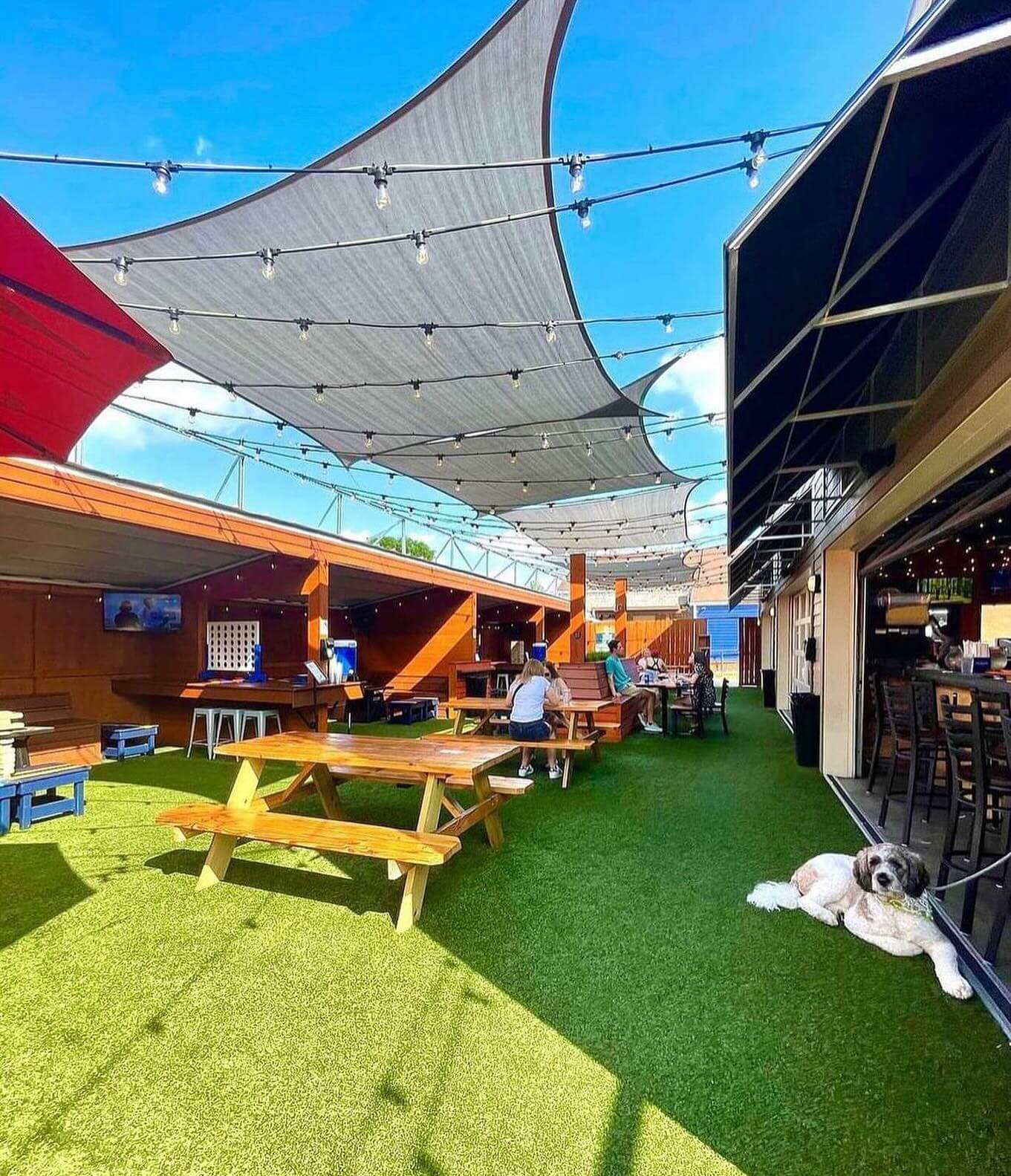 An outdoor seating area in Nashville featuring picnic tables, string lights, and shaded awnings with a person sitting and a dog lying on artificial grass.