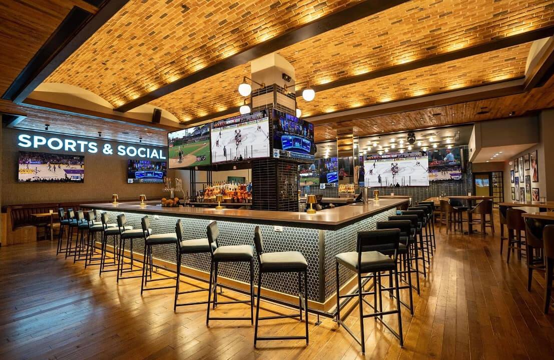 A spacious sports bar in Nashville with a central square-shaped bar area, multiple screens showing football games, bar stools, wooden floors and ceiling, and a sign reading "Sports & Social" on the wall.