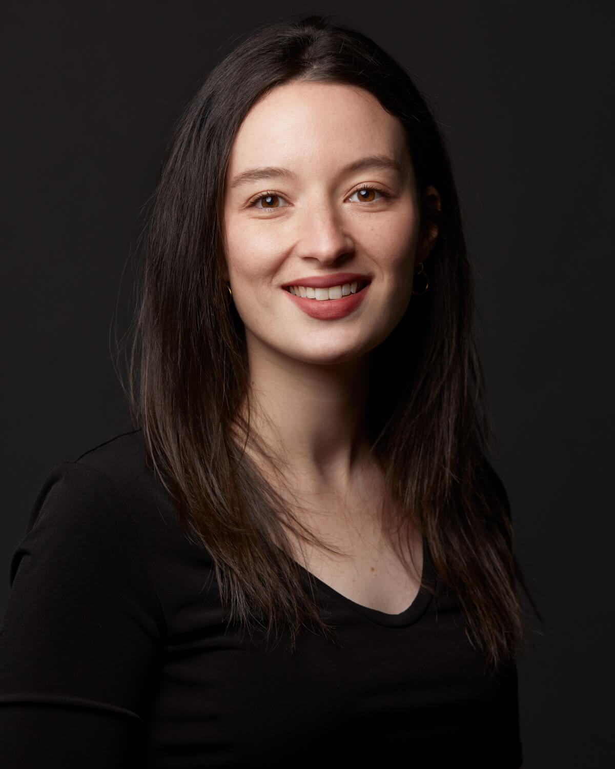 A woman with long brown hair wearing a black shirt, smiling subtly against a dark background.