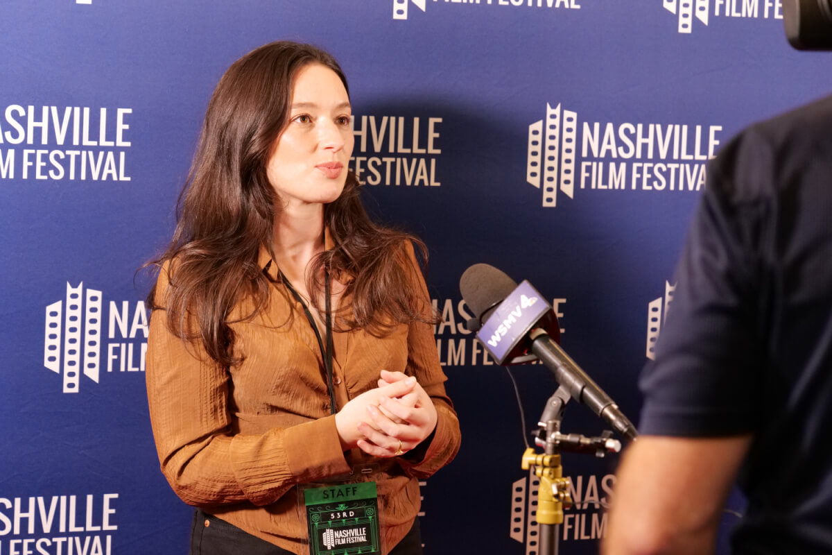 A woman is speaking into a microphone at the Nashville Film Festival, standing in front of a blue backdrop with the event's logo. She is wearing a brown shirt and a festival staff badge around her neck.